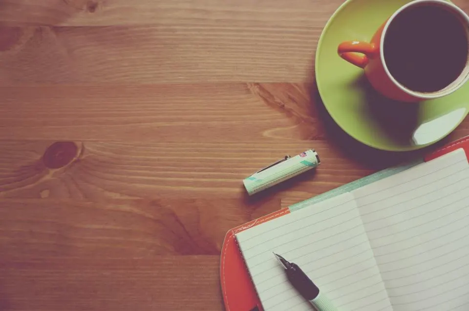 An open notebook, pen, and coffee cup on a wooden desk.